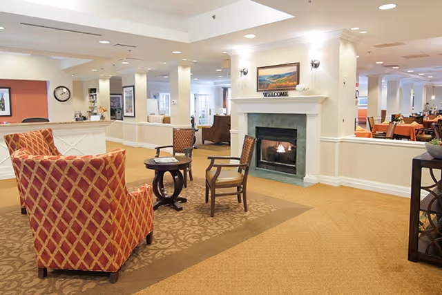 Communal lounge with armchairs and small tables arranged around a fireplace, with a dining area visible in the background.