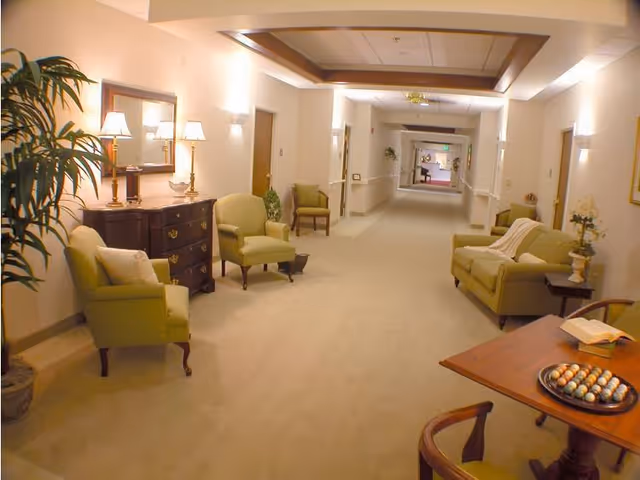 Well-lit interior hallway of a senior living community with green armchairs, side tables, and lamps.