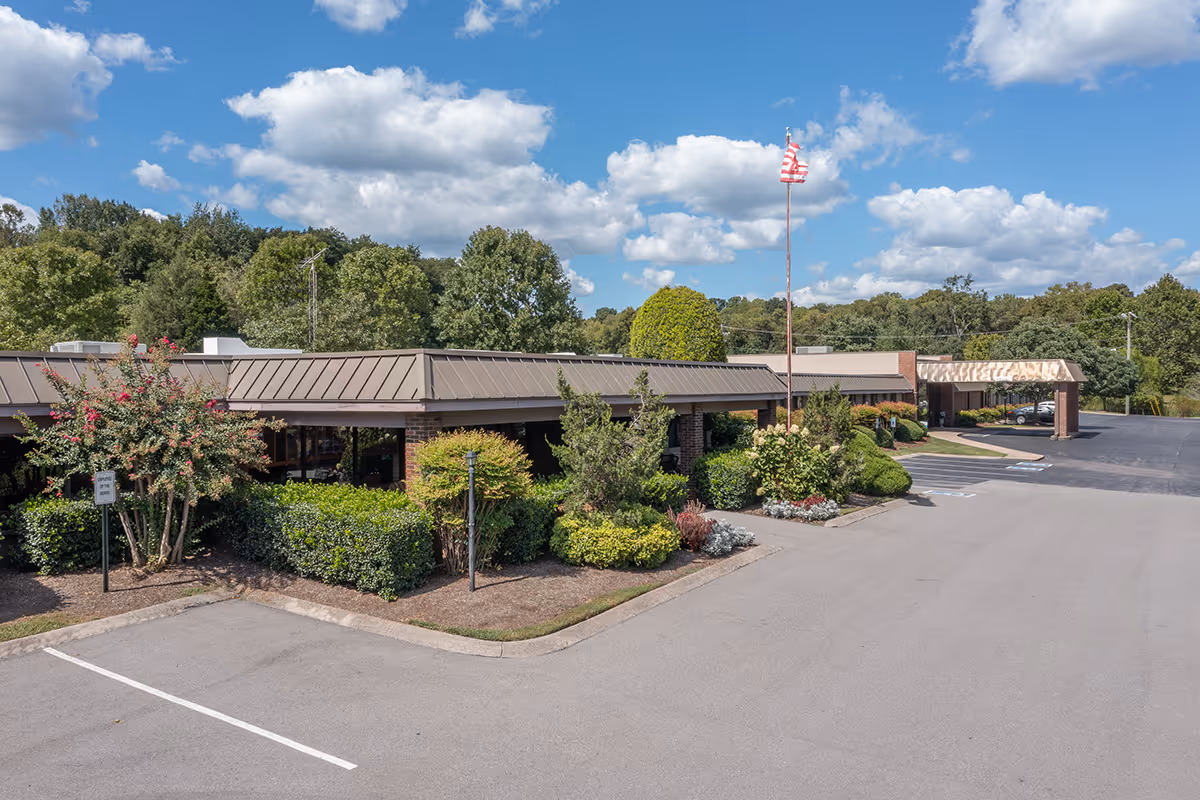 Low single-story senior living facility building with landscaped entrance, flagpole, and a mostly empty parking lot under a blue sky.