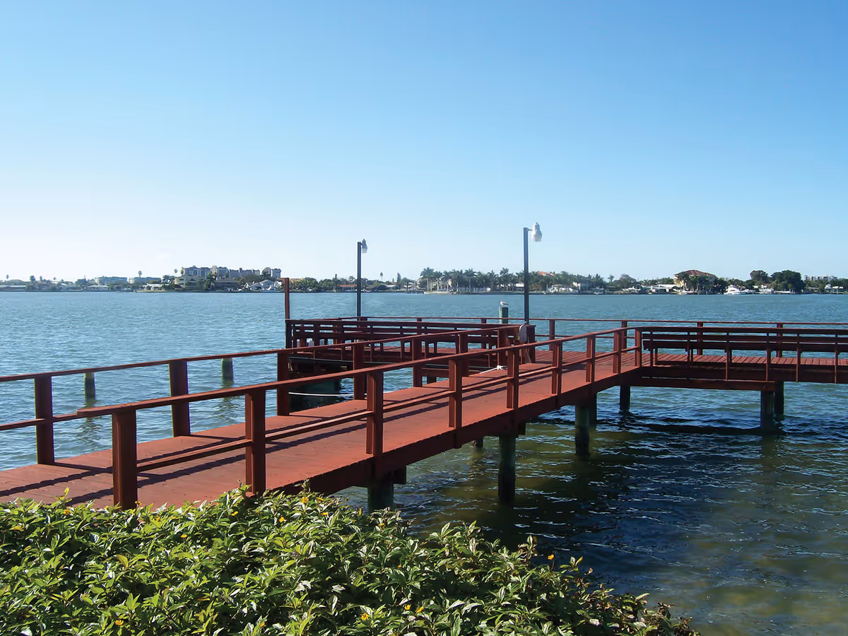 A wooden pier extending over a body of water with railings on both sides. There are two lamp posts on the pier and greenery in the foreground. The background shows a distant shoreline with buildings and trees under a clear blue sky.