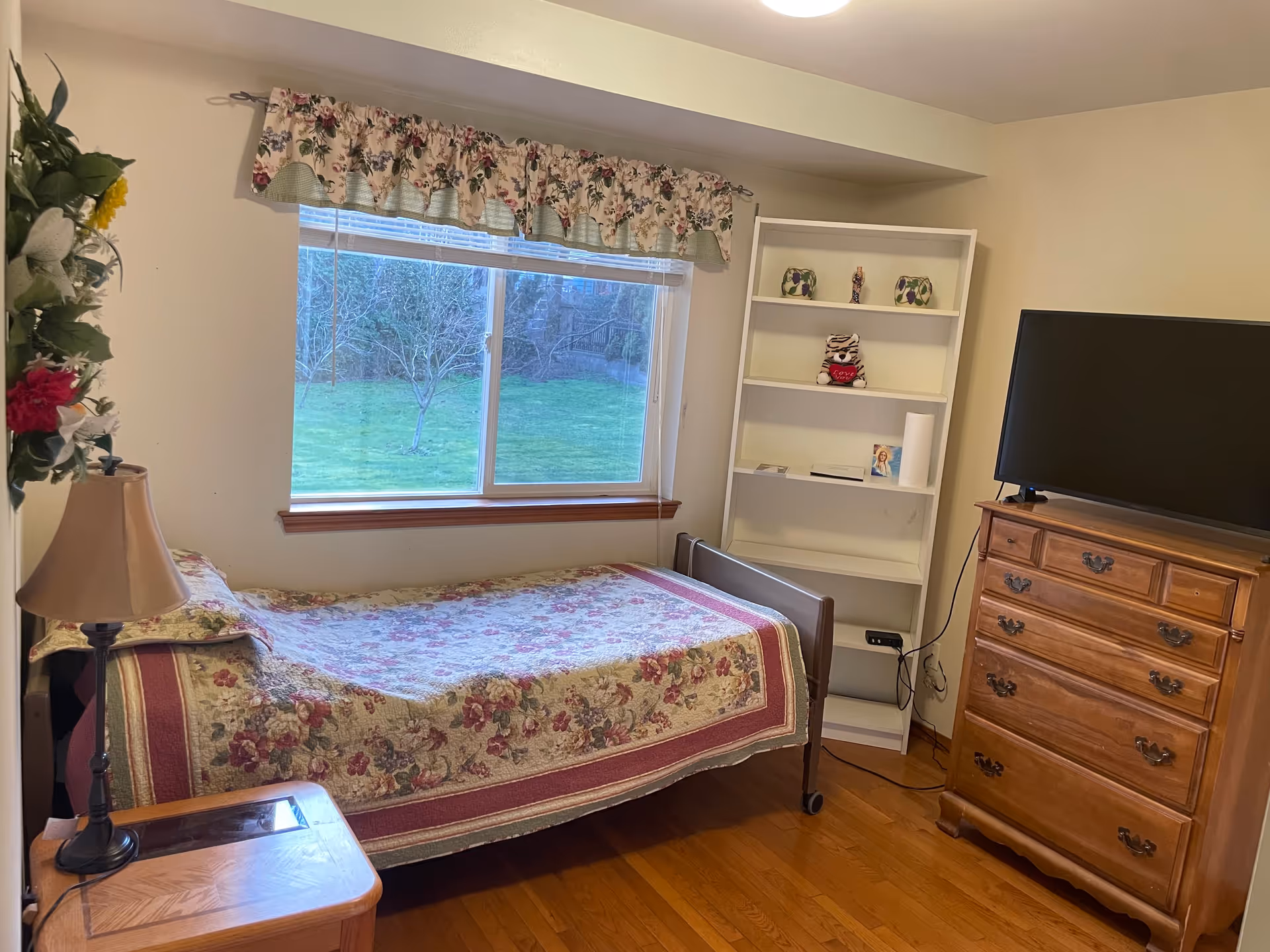 Small bedroom with a single bed covered in a floral quilt beneath a window, a white bookshelf, wooden dresser with a TV, and a bedside table with a lamp.