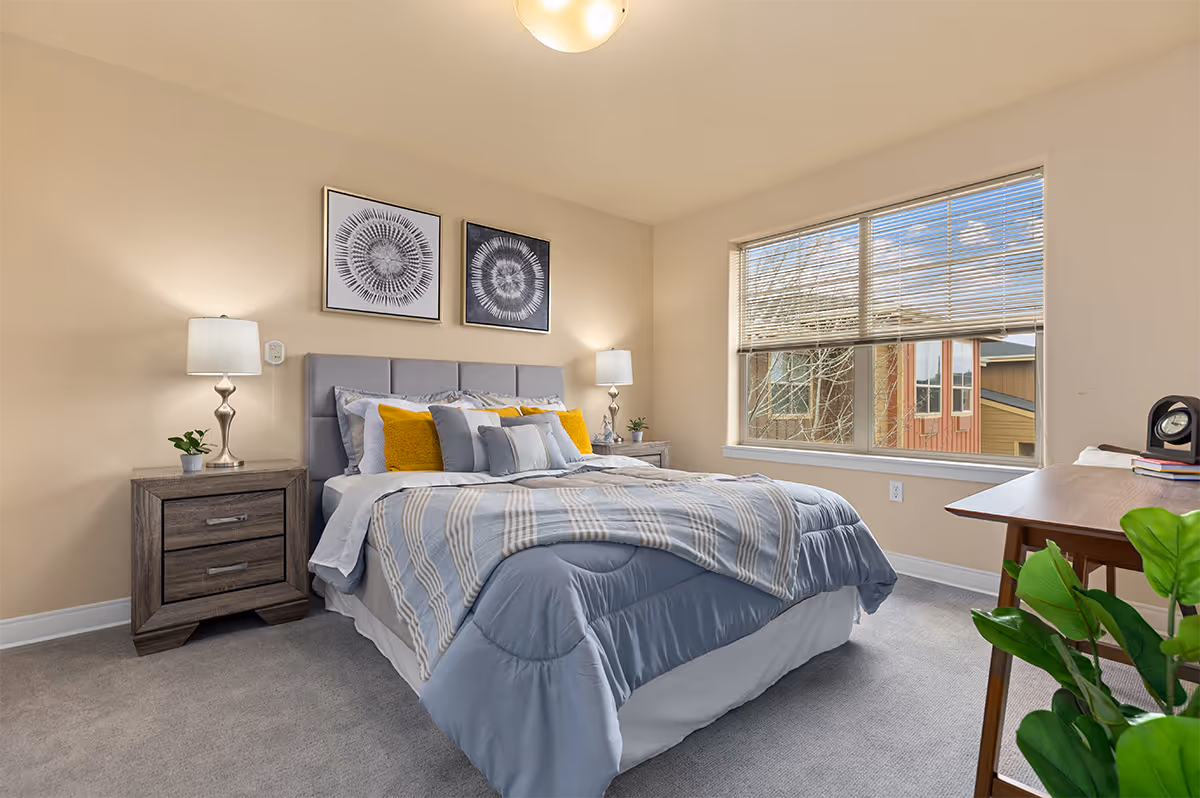 A neatly made bed with blue and white bedding and yellow accent pillows in a bright bedroom. The room has beige walls, two framed abstract artworks above the bed, two bedside tables with lamps and small plants, a large window with blinds partially open showing an outside view, a wooden desk with books and a clock, and a green plant in the foreground.