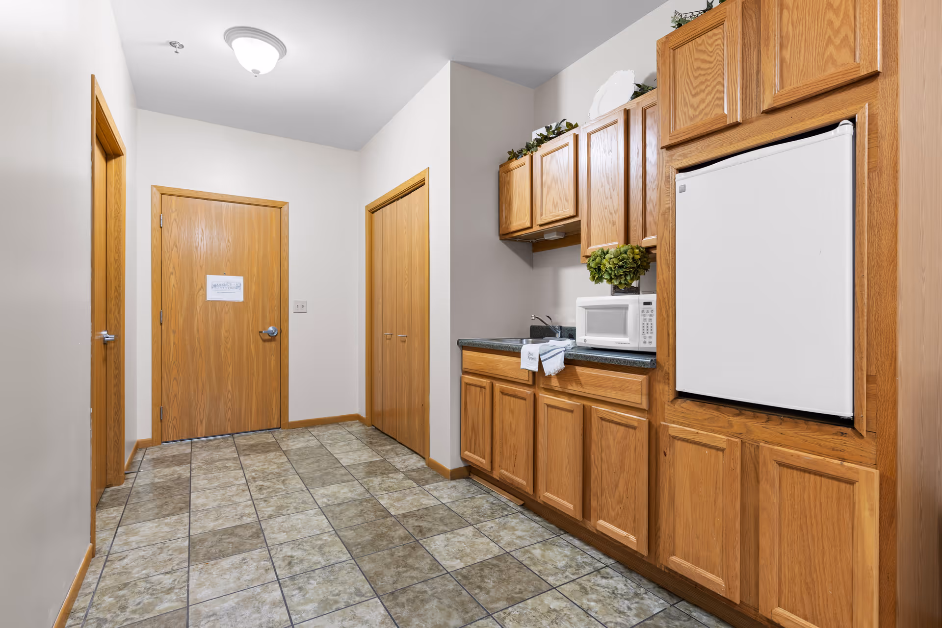Small kitchenette with oak cabinets, a microwave, mini-fridge, sink, and tiled floor in a hallway with three wooden doors.