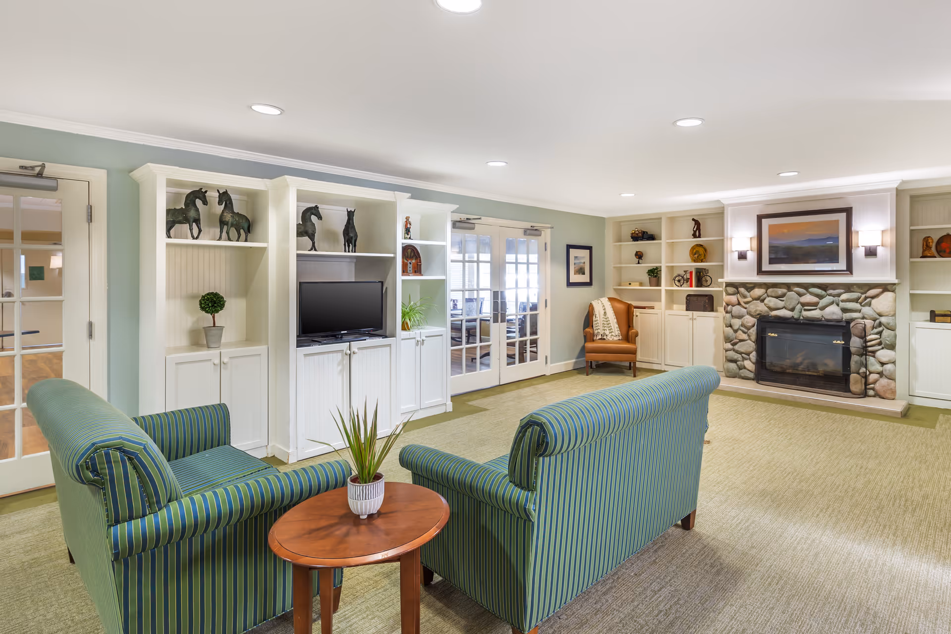 A cozy living room area in a senior living facility featuring two green striped armchairs facing a small wooden table with a potted plant. The room has built-in white shelves decorated with horse figurines, plants, and other decorative items. There is a stone fireplace with a framed painting above it, and a brown leather armchair with a throw blanket in the corner. French doors lead to another room with a conference table and chairs.