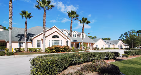 Front exterior of a single-story senior living building with palm trees, a driveway, and landscaped hedges under a clear blue sky.