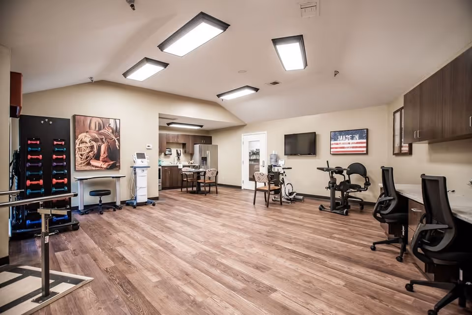 A spacious rehabilitation room with wooden flooring, exercise equipment including hand weights, stationary bikes, and parallel bars. There are chairs, a wall-mounted TV, and a kitchenette area with a table and chairs in the background. The room is well-lit with ceiling lights and has neutral-colored walls with framed artwork.