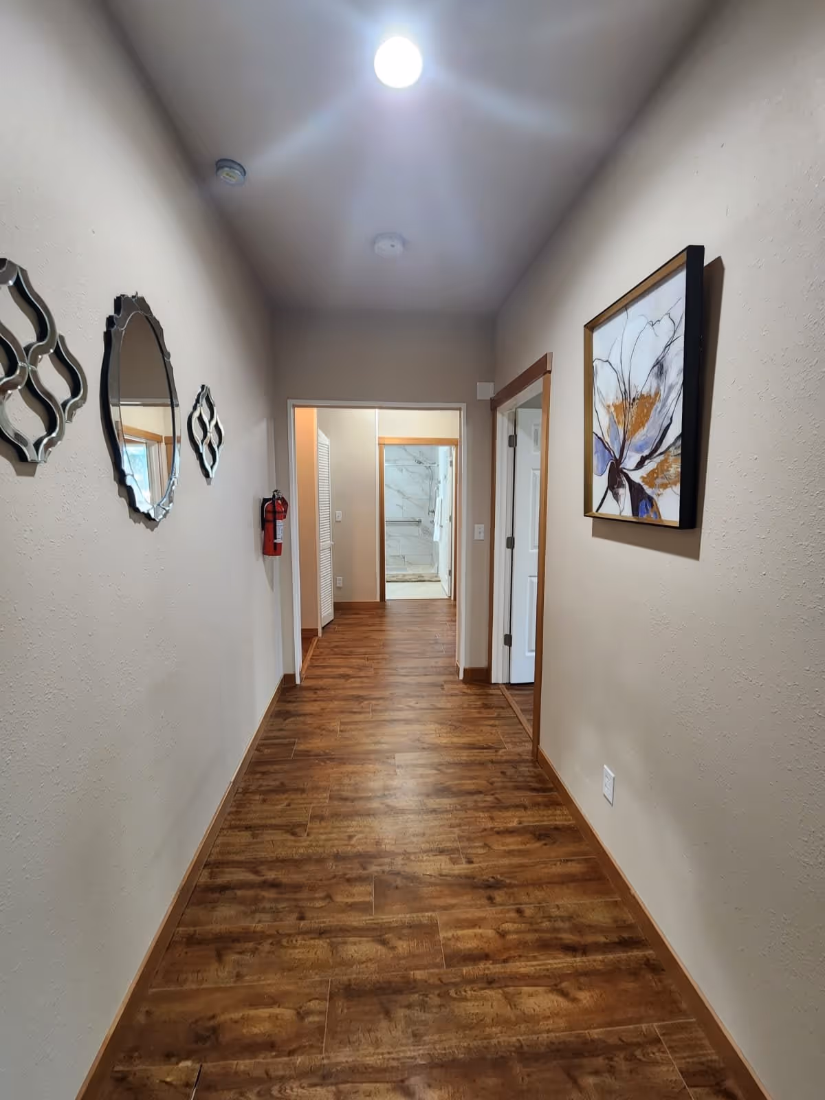 A hallway with wooden flooring and beige walls. On the left wall, there are three decorative mirrors and a fire extinguisher mounted. On the right wall, there is a framed floral artwork. At the end of the hallway, an open doorway reveals a bathroom with white marble tiles and a towel hanging on a rail.