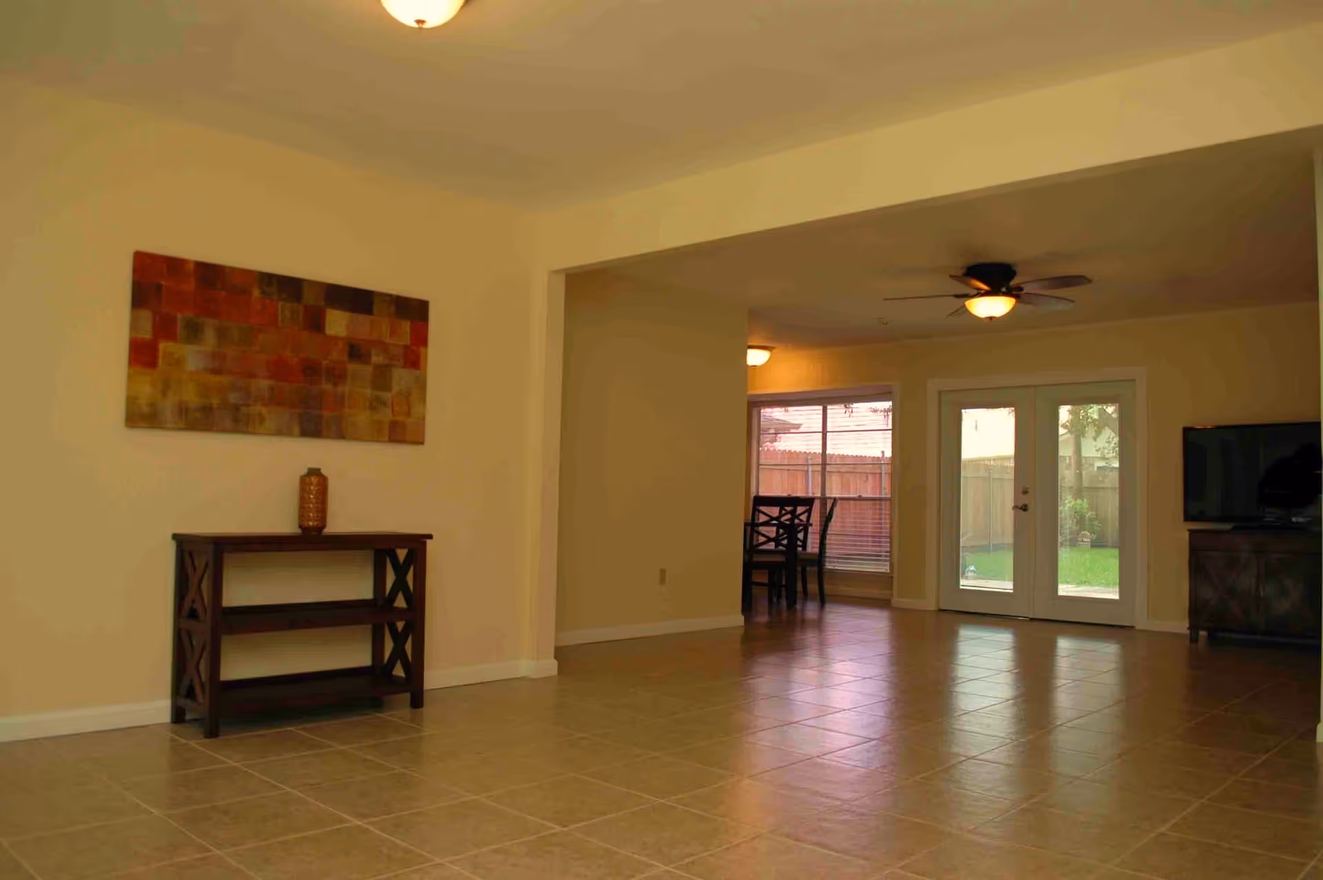Spacious tiled open living and dining area with a console table, wall art, ceiling fans, and glass doors to a backyard.