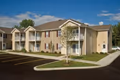 Two-story beige apartment building with balconies, a paved parking area, and small landscaped lawns under a blue sky.
