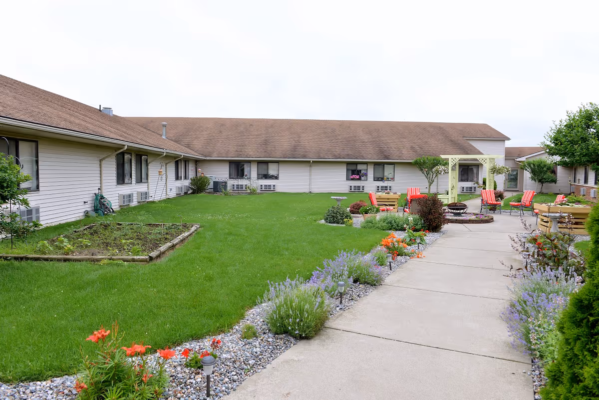Outdoor courtyard of a single-story senior living facility with a concrete walkway, green lawn, flowerbeds, and a seating area with red chairs.