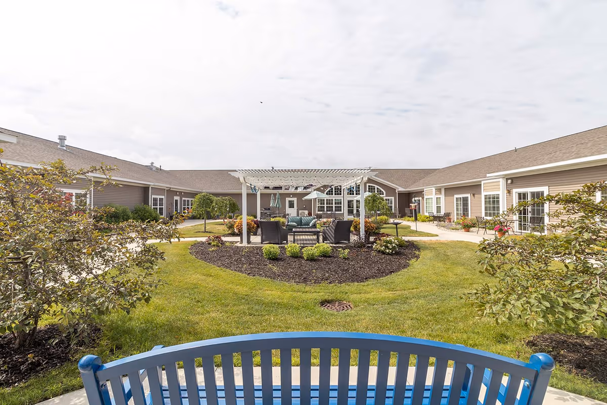 Outdoor courtyard area of Grand Village Assisted Living & Memory Care featuring a blue bench in the foreground, a circular garden with a white pergola and outdoor seating in the center, surrounded by well-maintained grass, shrubs, and a single-story building with multiple windows and doors.
