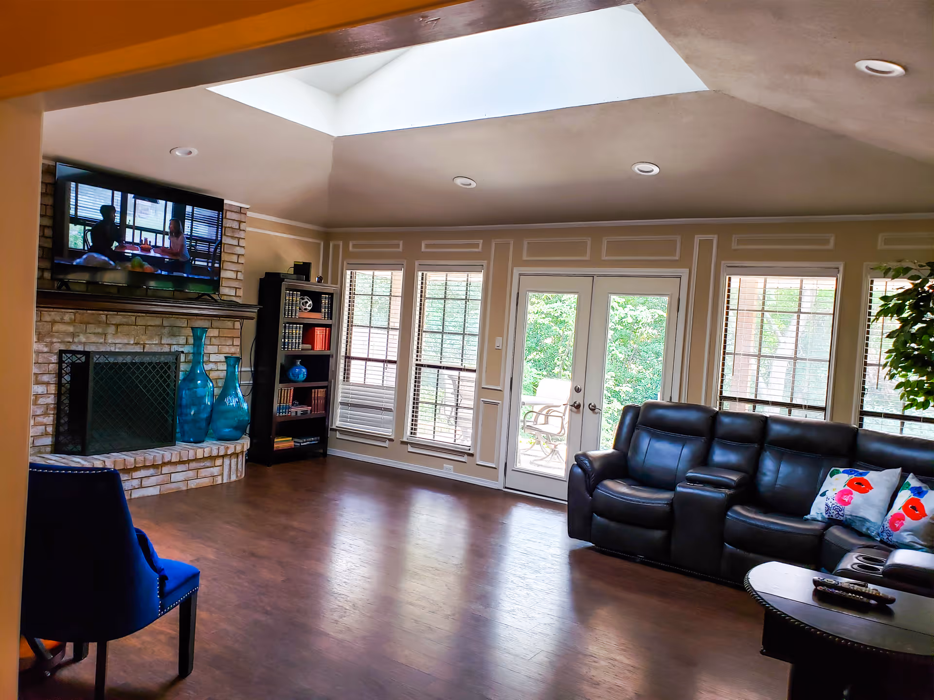 Bright living room with a leather reclining sofa, fireplace with TV above, large windows and a skylight.
