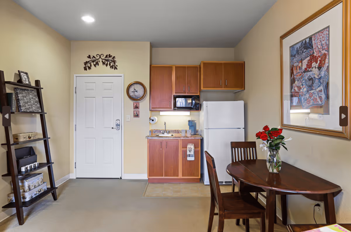 Interior view of a small kitchen and dining area in an assisted living facility. The kitchen features wooden cabinets, a white refrigerator, a microwave, a small sink, and a countertop with a coffee maker and cups. To the right, there is a wooden dining table with two chairs and a vase with red and white flowers. On the left side, there is a dark wooden ladder shelf with decorative items. The walls are beige, and there is a framed colorful painting hanging above the dining table.