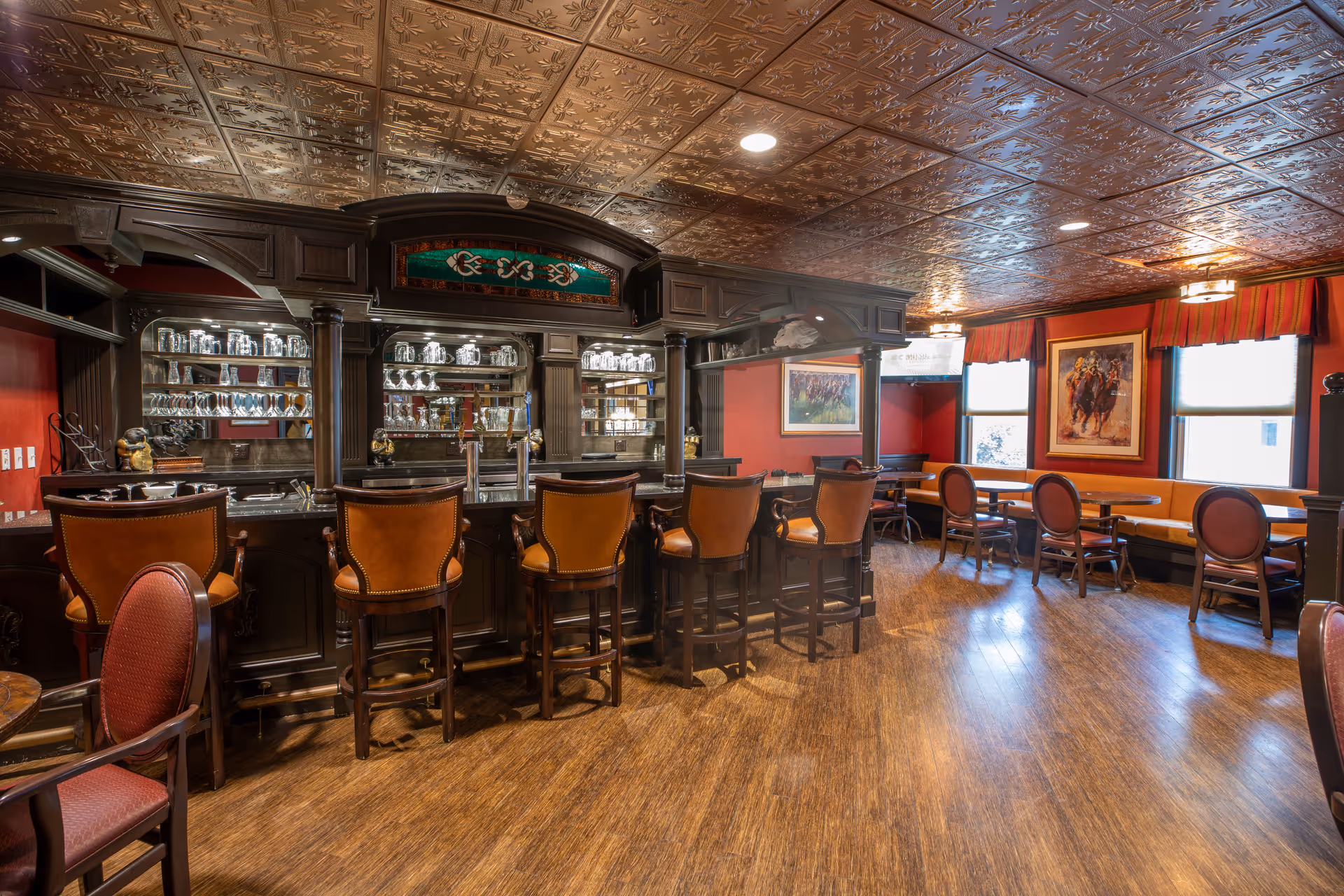 Interior view of a senior living facility's bar and lounge area with a dark wood bar counter, leather bar stools, and glassware displayed on shelves behind the bar. The room features a decorative tin ceiling, wooden flooring, red walls, framed artwork, and seating areas with tables and chairs near windows with red and orange striped valances.
