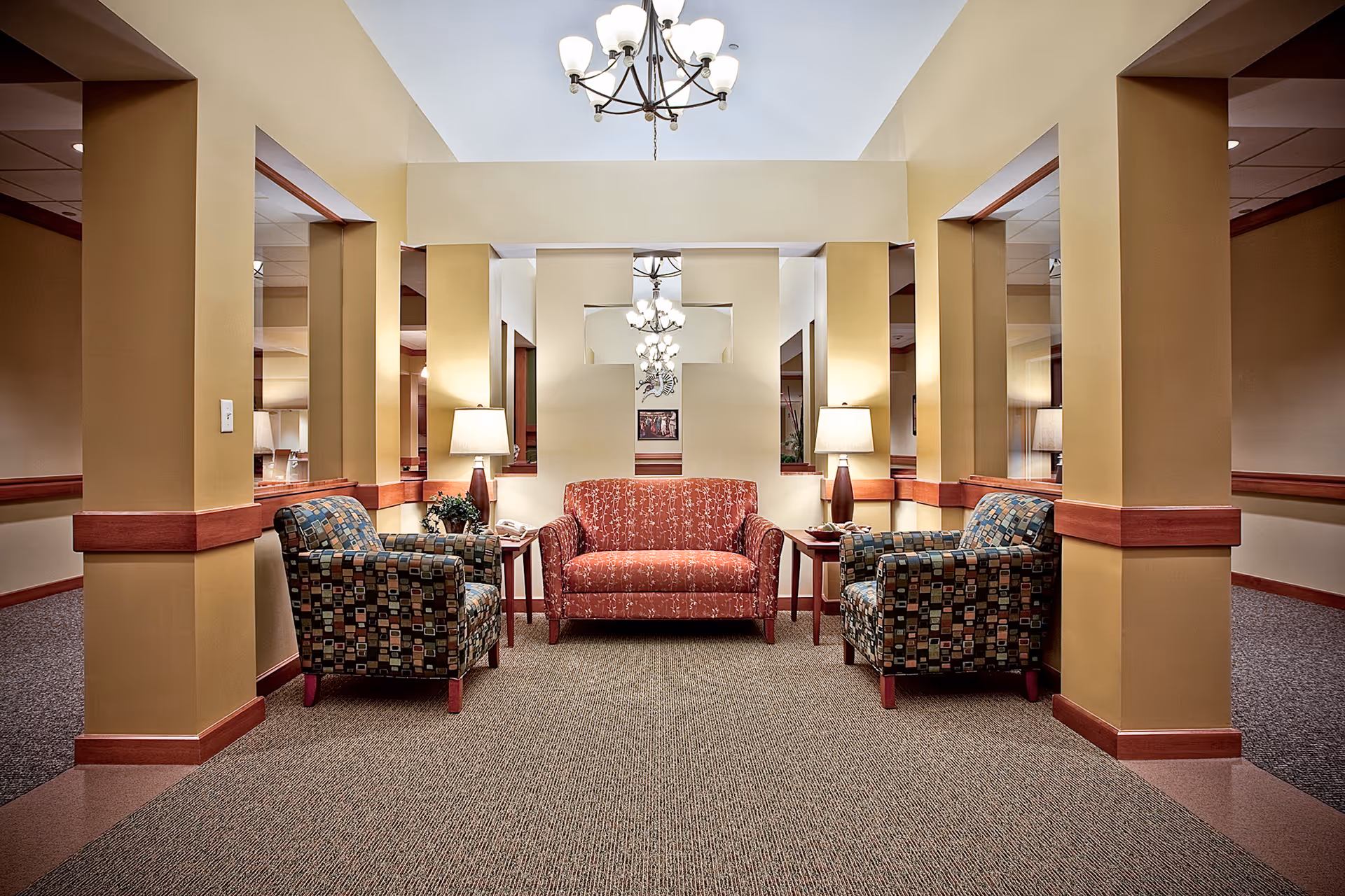 A cozy seating area in a senior living facility with a red patterned loveseat flanked by two multicolored patterned armchairs. Each side of the loveseat has a wooden side table with a lamp. The walls are painted beige with wooden trim, and there are multiple mirrors and a chandelier overhead, creating a warm and inviting atmosphere.