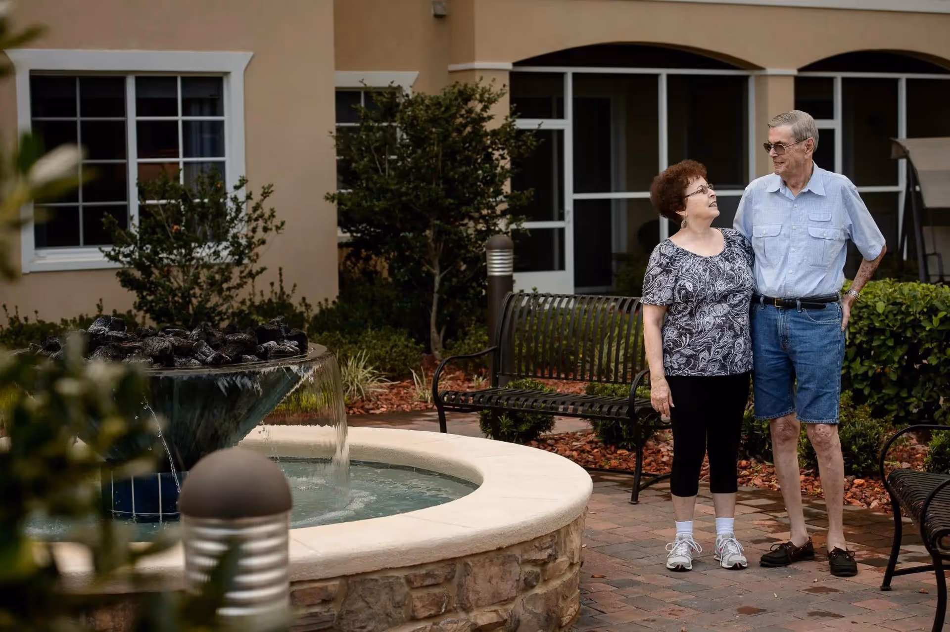 Two older adults stand in a courtyard by a round fountain with benches and a building facade behind them.