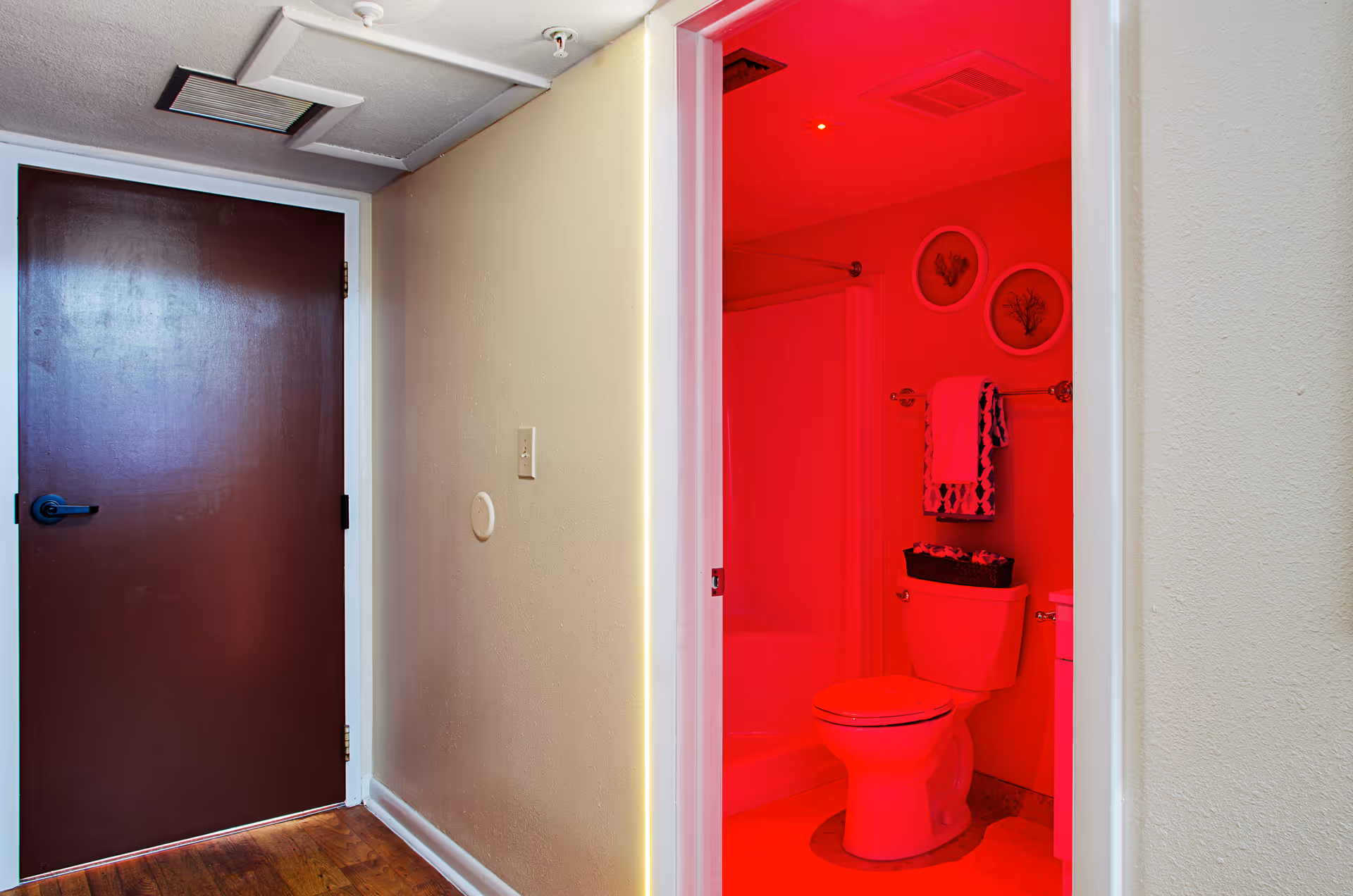 View of a hallway with a closed dark brown door on the left and an open door on the right revealing a bathroom with a toilet, towel rack with towels, and framed wall art. The bathroom is illuminated with red lighting.