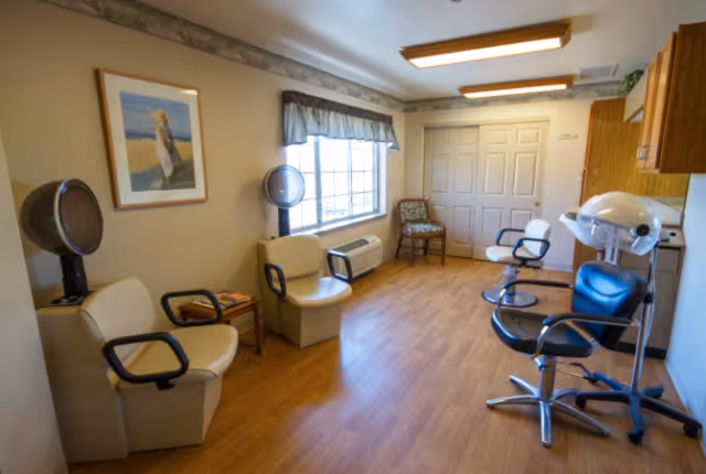A small salon room with wooden flooring and beige walls. The room contains two beige salon chairs with hair dryers attached, two black salon chairs, a wooden chair with a patterned cushion, a small wooden side table with magazines, a framed picture on the wall, and a window with a valance letting in natural light.
