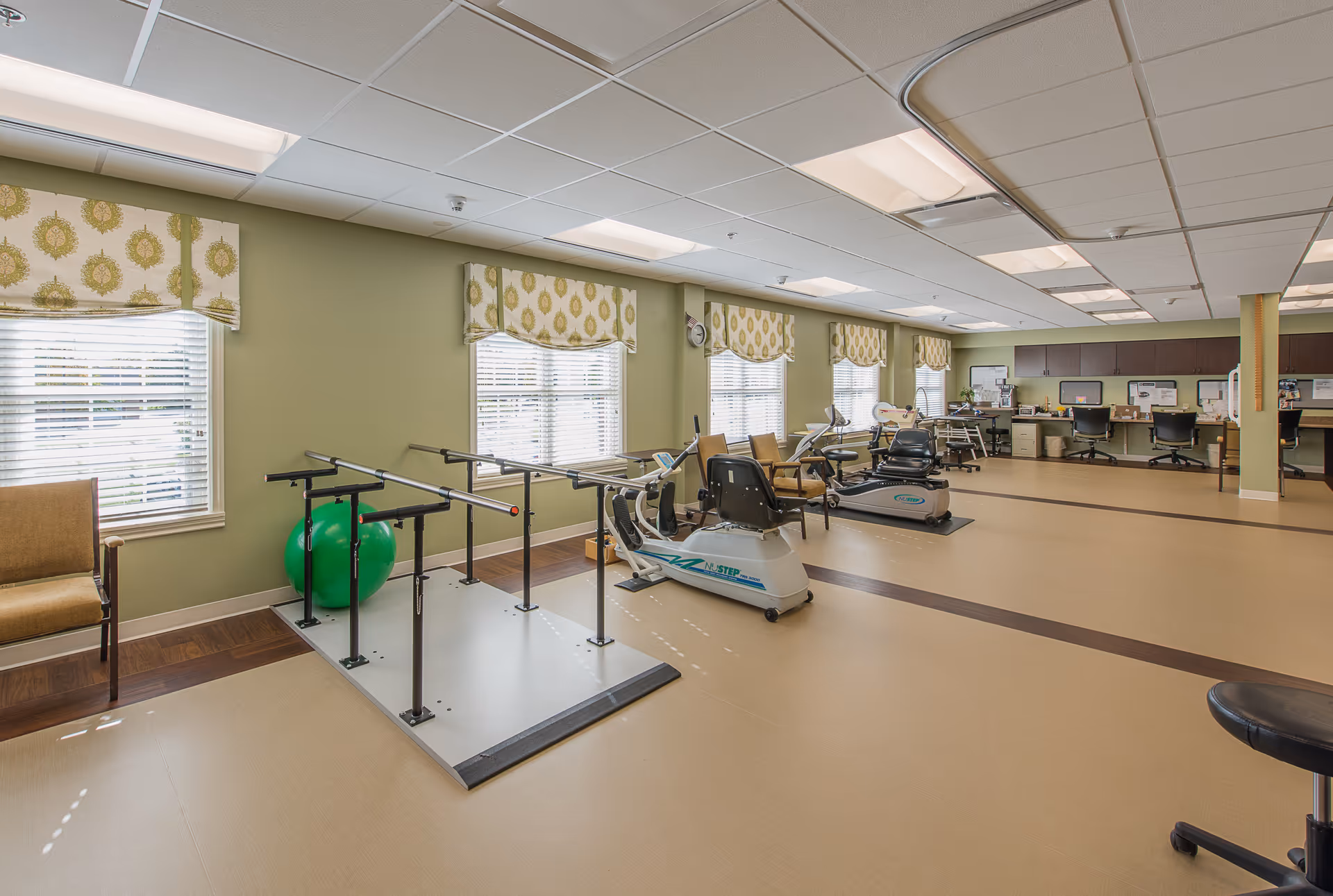 A spacious interior therapy/exercise room with parallel bars, recumbent exercise bikes, chairs and workstations under ceiling lights.