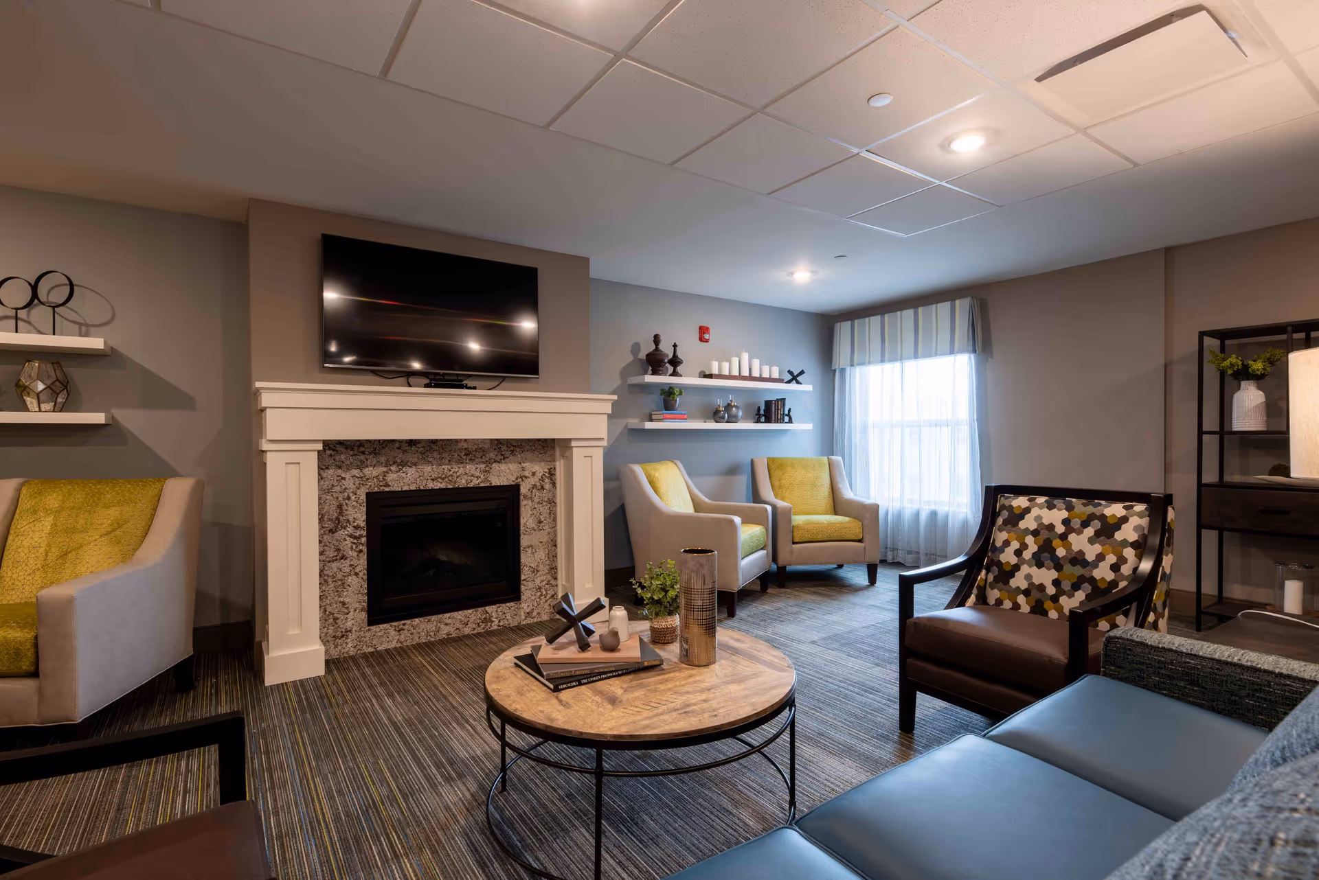 A cozy living room with a modern fireplace and a mounted flat-screen TV above it. The room features a round wooden coffee table with decorative items, several armchairs with green and patterned cushions, a blue sofa, and shelves with books and decorative objects. A window with sheer curtains allows natural light into the space.