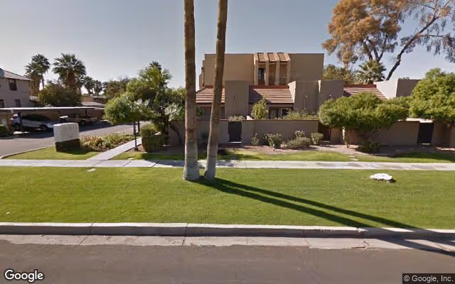 Exterior view of a single-story senior living facility building with a flat roof and beige walls, surrounded by green grass, palm trees, and shrubs under a clear sky.