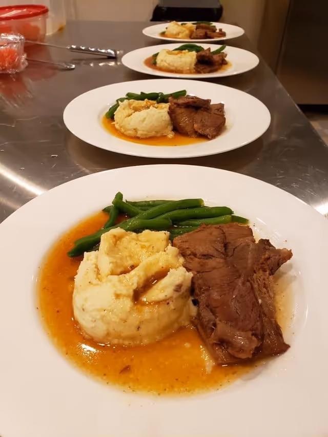 Plated meals of roast beef, mashed potatoes with gravy, and green beans lined up on a stainless counter.