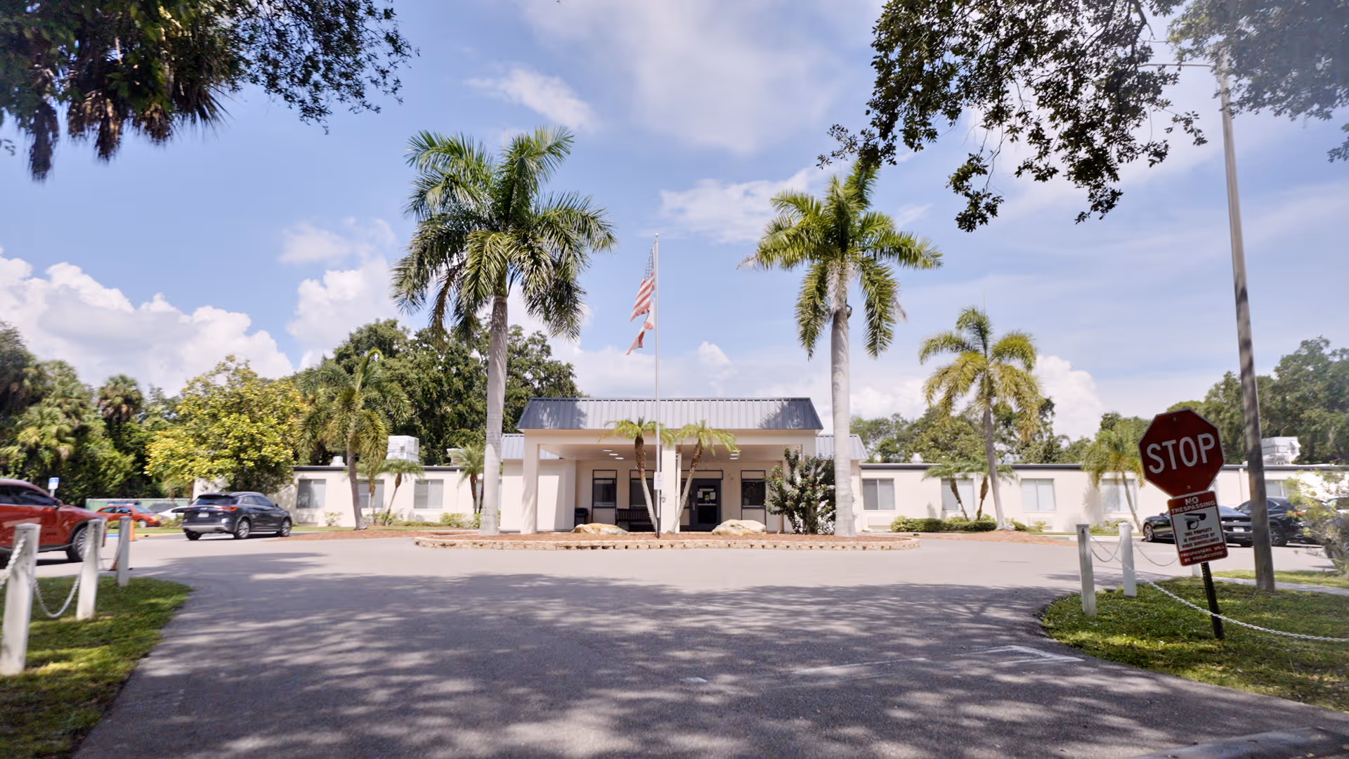 Front exterior view of Woodbridge Care Center building with palm trees, a driveway, parked cars, and a stop sign under a partly cloudy sky.