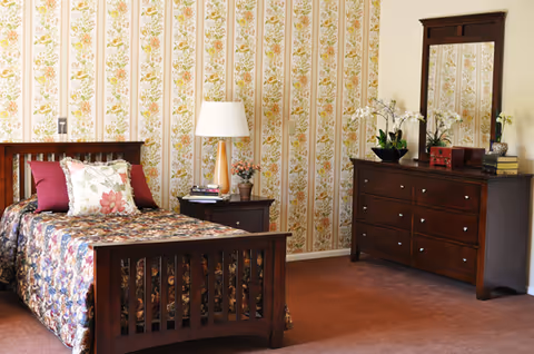 A cozy bedroom with a wooden bed frame, floral bedspread, and decorative pillows. Next to the bed is a wooden nightstand with a lamp, books, and a small plant. Across from the bed is a wooden dresser with a mirror, adorned with potted orchids and decorative items. The walls have floral patterned wallpaper and the floor is carpeted.