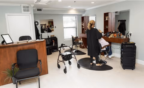 Interior view of a salon area in a senior living facility with a stylist cutting a seated customer's hair. The room has light-colored walls, a large window, salon chairs, mirrors, and various hair styling tools. A walker is visible near the center of the room.