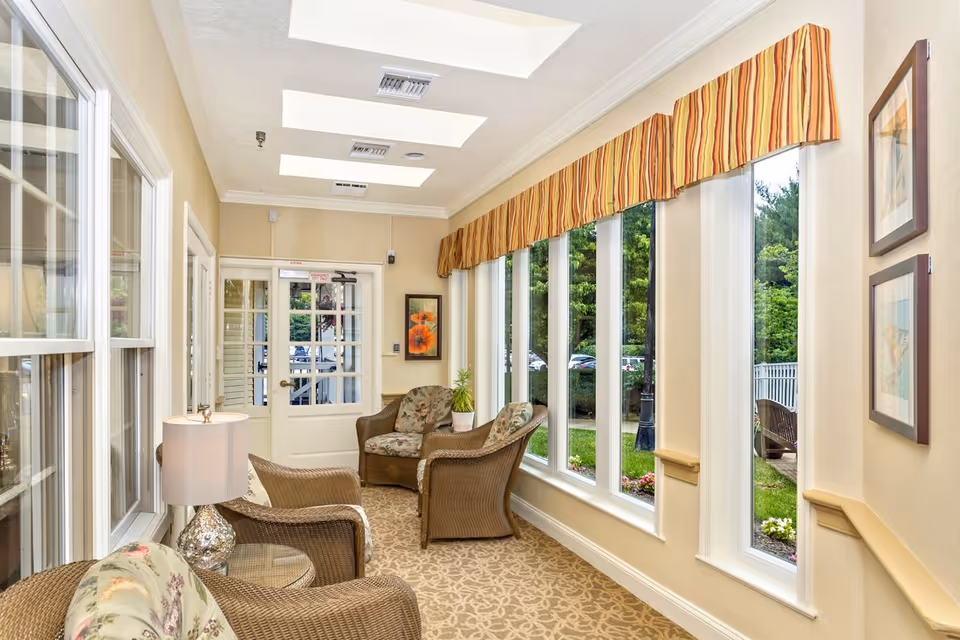 Bright sunroom-style sitting area with wicker chairs, a lamp, and large windows overlooking a garden.