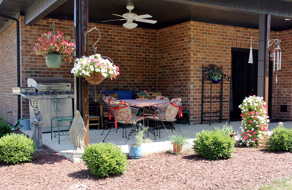 Outdoor patio area with a brick wall background featuring a round table with four cushioned chairs, a ceiling fan, hanging flower baskets, a stainless steel grill, garden statues, and various potted plants surrounded by mulch and small bushes.