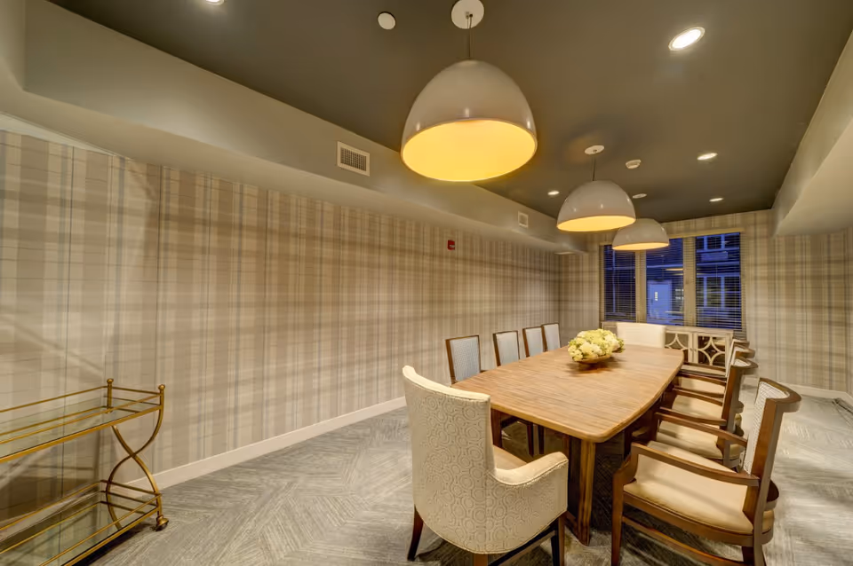 A well-lit dining room with a long wooden table surrounded by eight upholstered chairs. The walls have a beige plaid pattern, and three large pendant lights hang from the ceiling above the table. A decorative floral centerpiece is placed on the table, and there is a window with blinds at the far end of the room. A small gold-colored serving cart is positioned against the left wall.