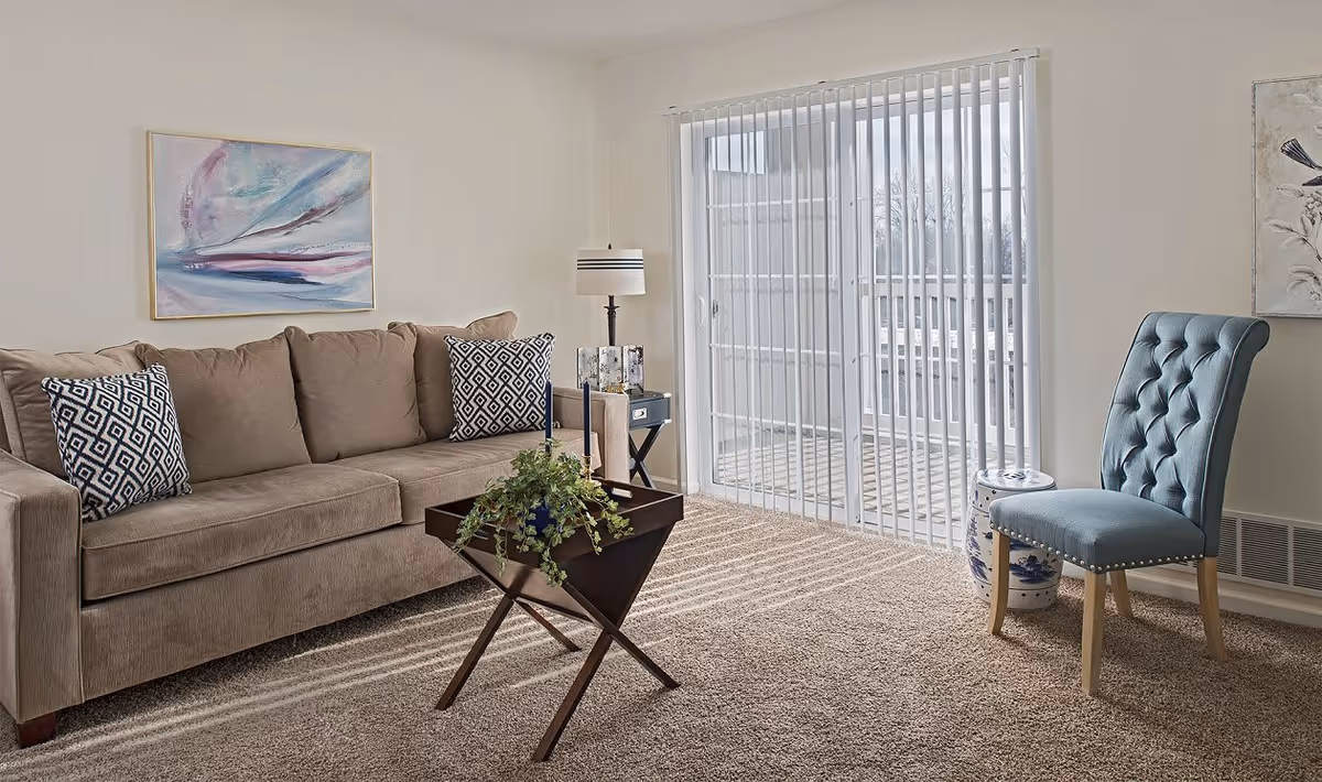 Sunlit living room with a beige sofa, patterned pillows, a small wooden coffee table with a plant, and a blue tufted chair by sliding glass doors.