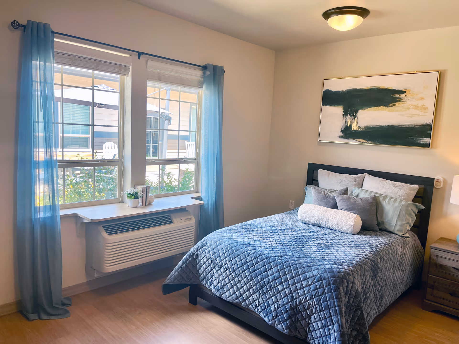 Sunlit bedroom with a neatly made bed covered in a blue quilt, a window with teal curtains above an air-conditioning unit, wall artwork and a nightstand.