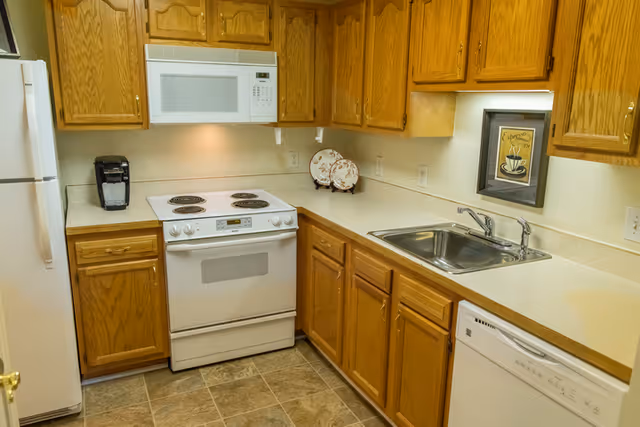 A compact kitchen with wooden cabinets, a white refrigerator, a white microwave mounted above a white electric stove, a coffee maker on the counter, a double sink with a faucet, a dishwasher, and decorative plates and a framed coffee-themed picture on the wall.
