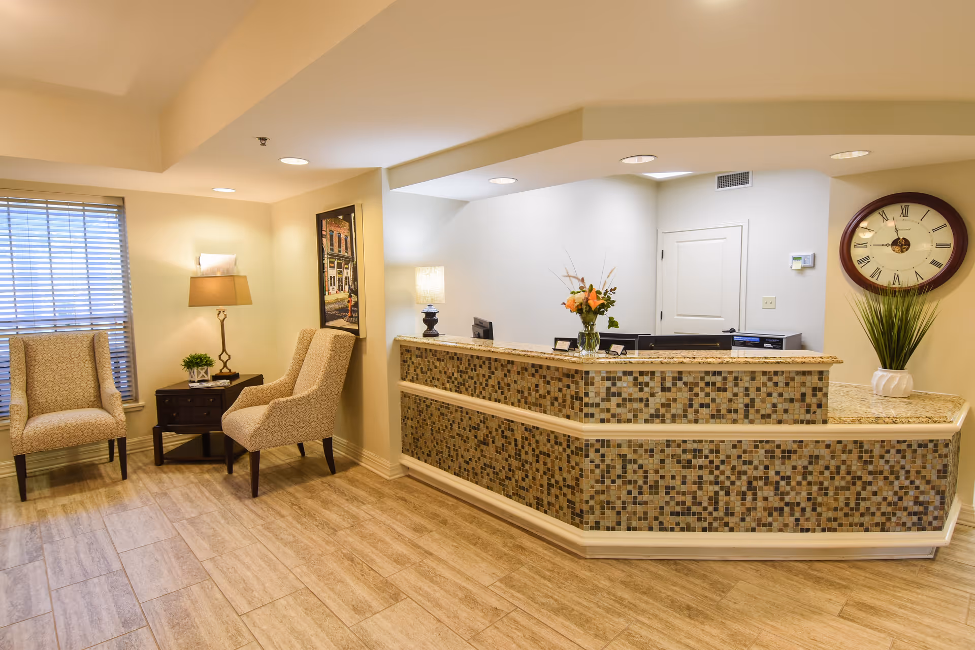 Welcoming reception area with a mosaic-tiled front desk, two upholstered chairs by a side table and lamp, and a wall clock.