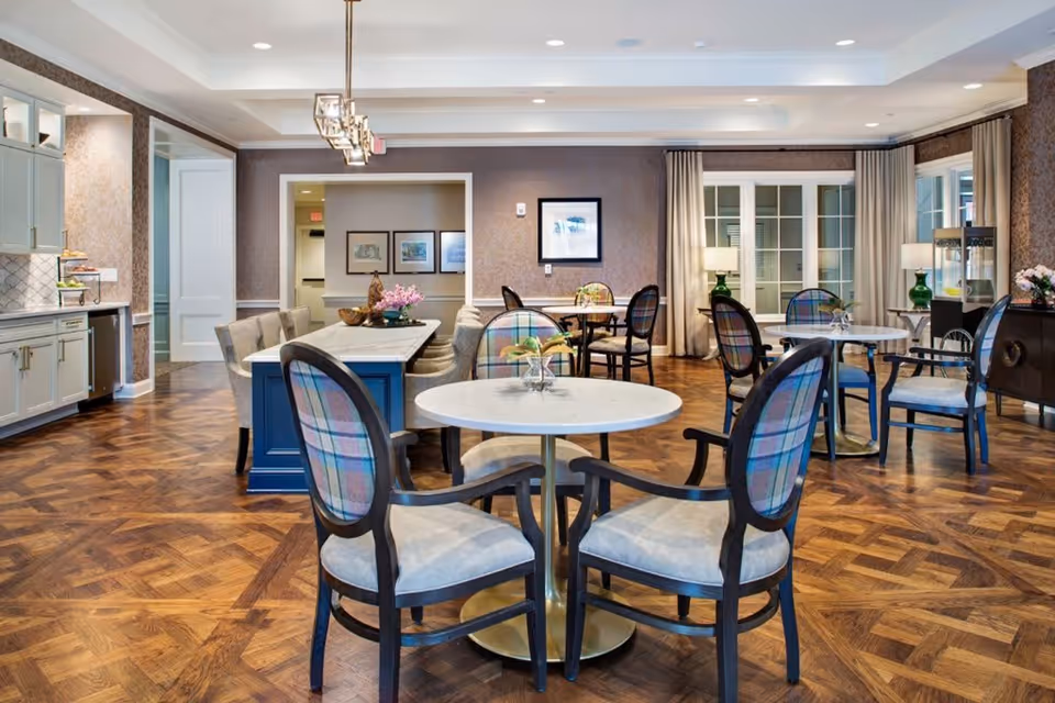 A spacious and elegant dining room with multiple round and rectangular tables surrounded by chairs with plaid upholstery. The room features wooden parquet flooring, soft lighting from ceiling fixtures, and large windows with curtains. There is a sideboard with a lamp and decorative items, and framed artwork on the walls.