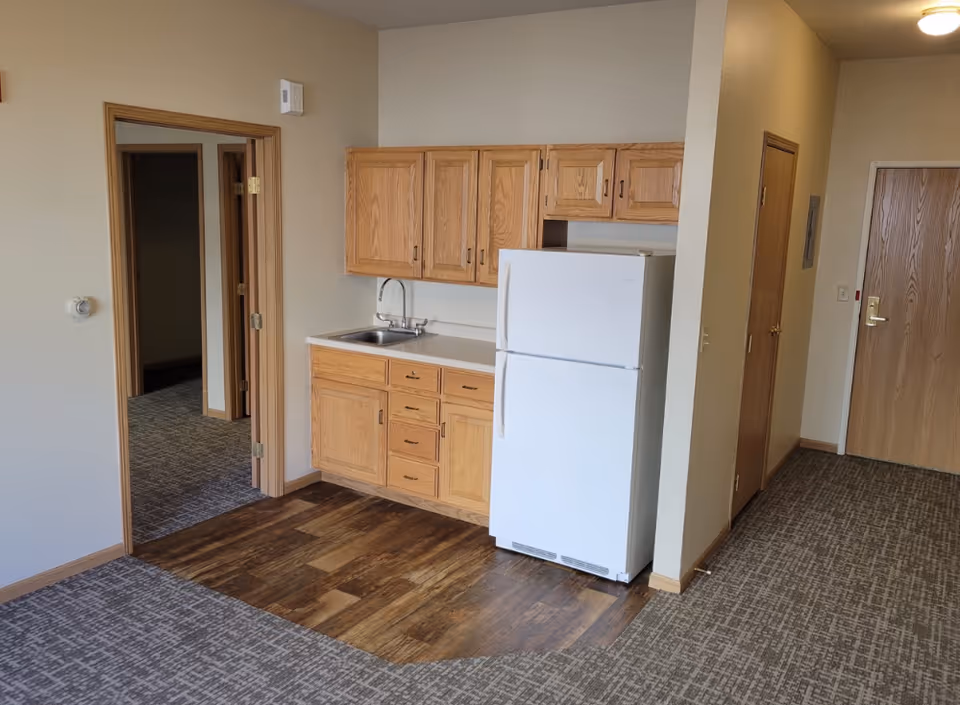 Interior view of a senior living facility unit showing a small kitchen area with wooden cabinets, a white refrigerator, and a sink. The floor transitions from carpet to wood-like flooring in the kitchen area. There is a doorway leading to another room and a hallway with a closed wooden door.