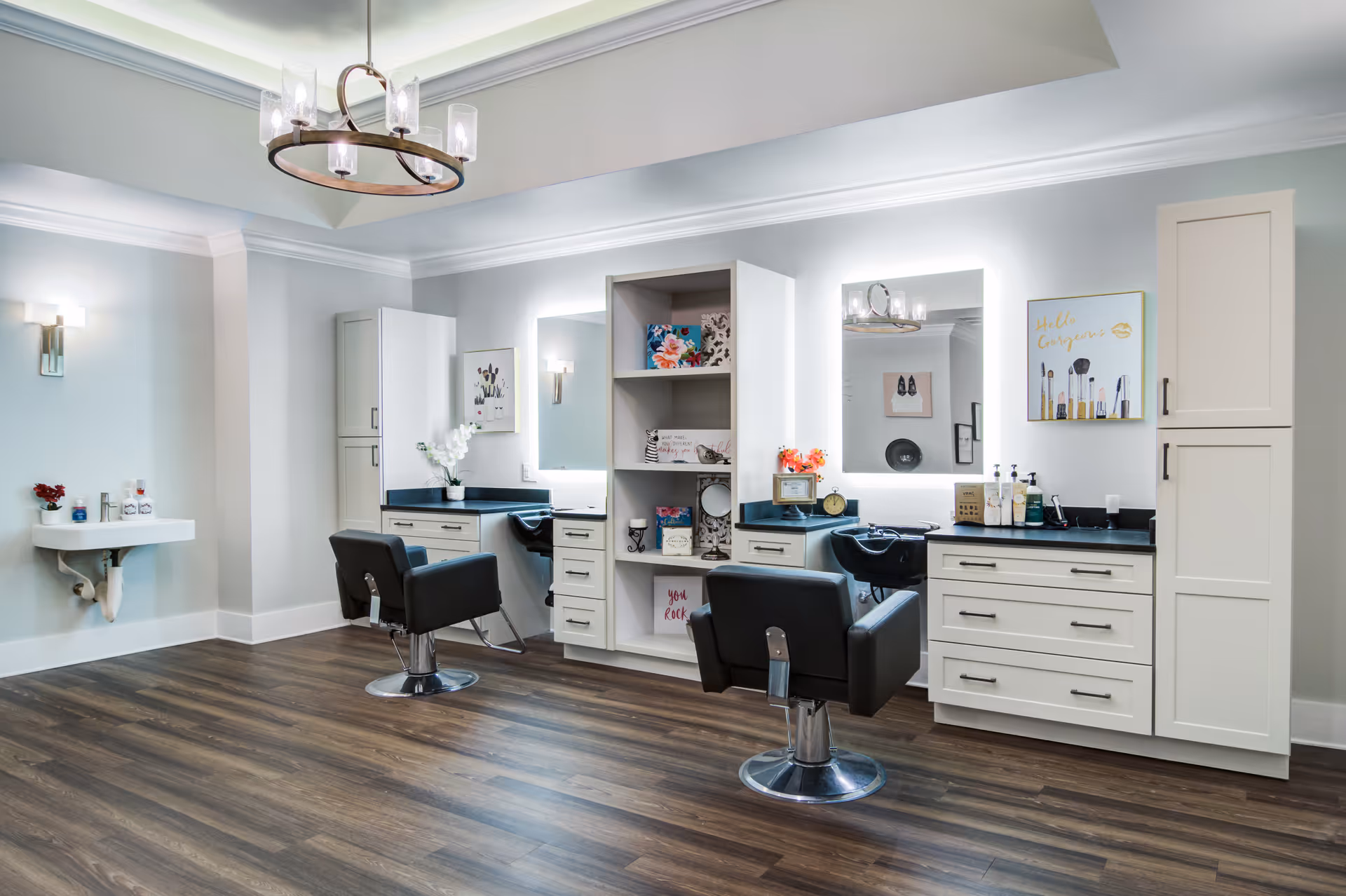 A modern hair salon area with two black salon chairs in front of two illuminated mirrors. The space features white cabinetry with drawers and shelves holding decorative items and hair products. There is a small white sink mounted on the wall to the left and a stylish chandelier hanging from the ceiling. The floor is wood with a dark finish.