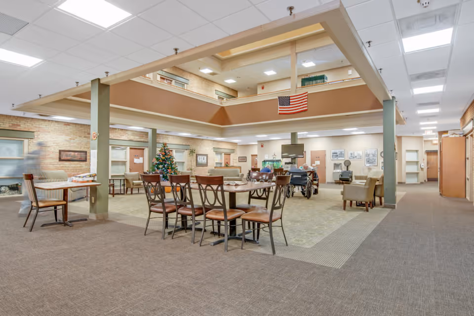 Open communal dining and lounge area in a senior living facility with tables, chairs, a TV, a Christmas tree, and an American flag on an upper balcony.