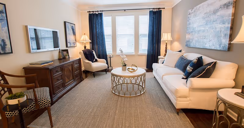 A cozy living room with a white sofa adorned with blue and white pillows, a round coffee table with decorative items, two armchairs, a wooden sideboard with books and picture frames, two standing lamps, and large windows with dark blue curtains letting in natural light.