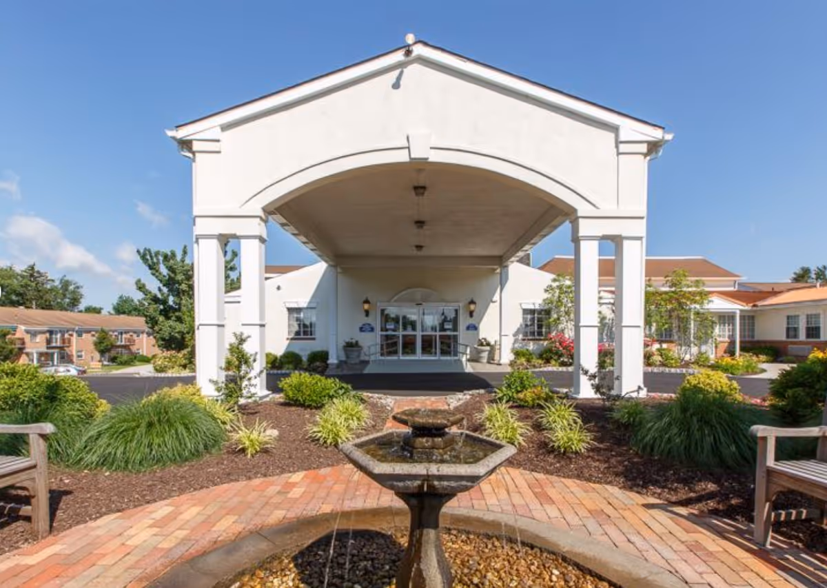 Entrance of Fort Washington Estates senior living facility with a covered driveway, landscaped garden with bushes and flowers, a water fountain in the foreground, and benches on either side.