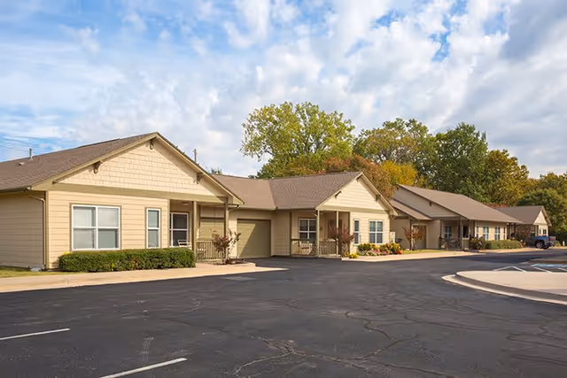 Exterior view of a single-story senior living facility building with beige siding, multiple entrances, small porches, and a paved parking lot in front. Trees with green and autumn-colored leaves are visible in the background under a partly cloudy sky.