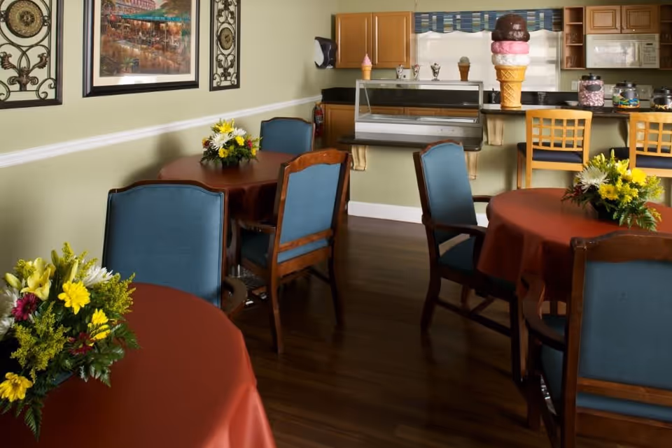 Interior view of a dining area with round tables covered in red tablecloths, each adorned with a floral centerpiece. Blue cushioned wooden chairs surround the tables. In the background, there is a counter with bar stools and decorative ice cream cone sculptures, along with wooden cabinets and a window with a blue valance.