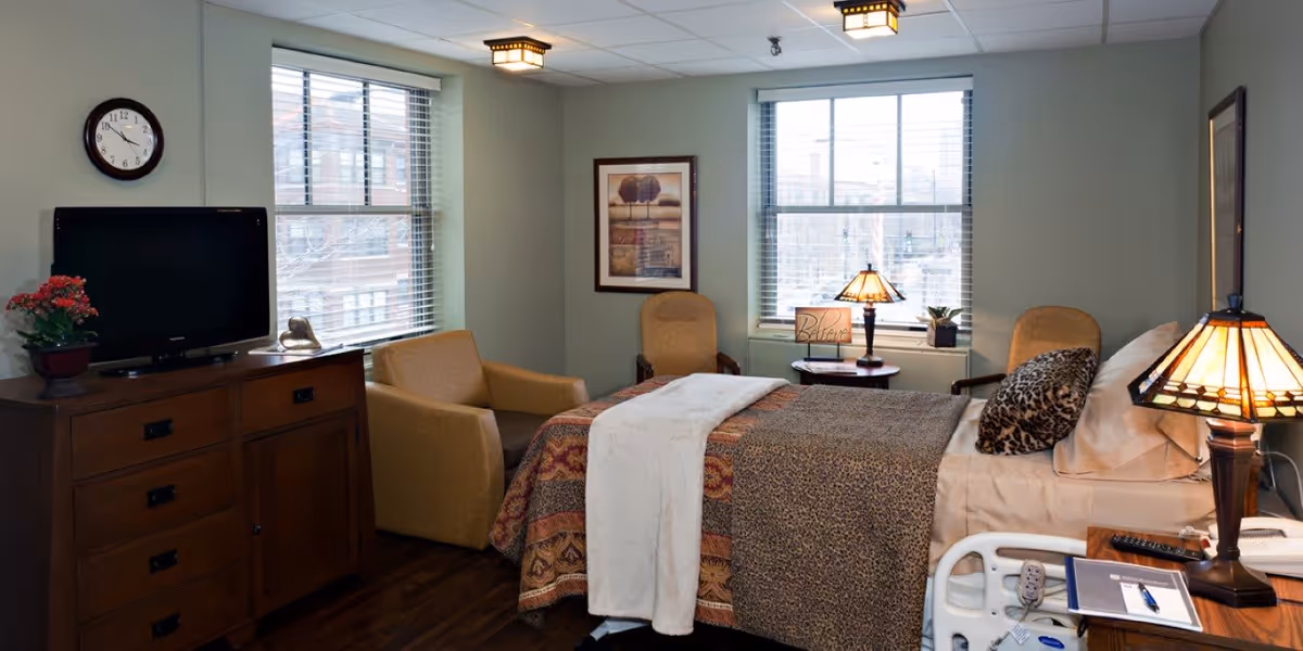A cozy bedroom in a senior living facility with a single bed covered in patterned blankets and a white throw. The room has two windows with blinds, a wooden dresser with a TV and a potted plant, two armchairs, a side table with a lamp and a decorative sign that says 'Believe'. There is also a bedside table with a lamp, a remote control, and a notepad with a pen.