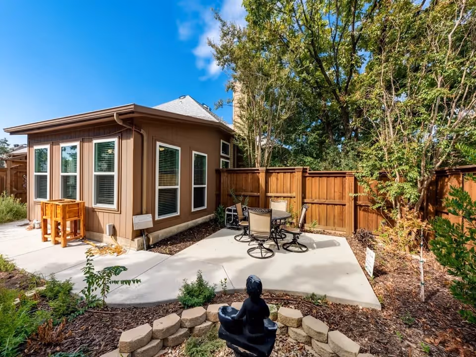 Outdoor patio area at Greenfield Residential Care featuring a concrete seating area with a round table and four chairs, surrounded by a wooden fence and lush green trees. In the foreground, there is a small garden with a black statue and stone border, under a bright blue sky with some clouds.