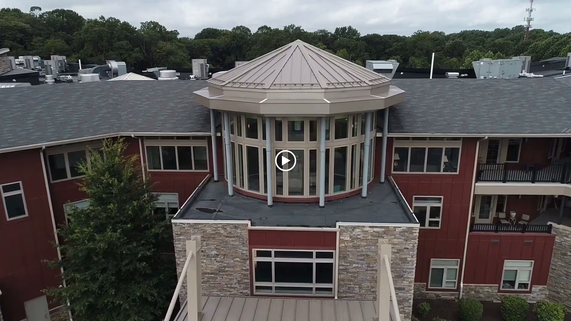 Exterior view of a senior living facility named The Summit, showing a multi-story building with a distinctive round turret-like structure with large windows and a conical roof. The building has red siding, stone accents, and multiple windows. Trees and greenery are visible in the background under a cloudy sky.