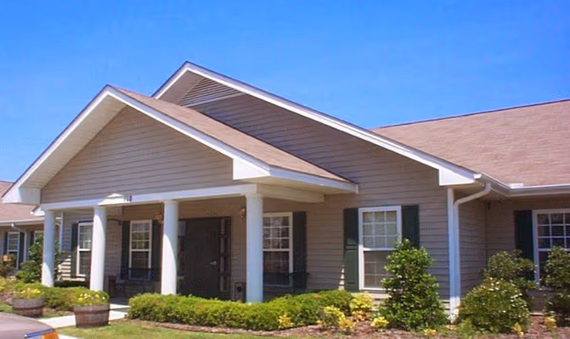 Exterior view of a single-story building with beige siding, white columns supporting a covered entrance, green window shutters, and a well-maintained garden with shrubs and flowers under a clear blue sky.