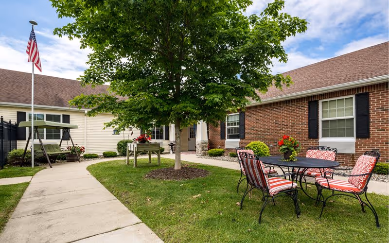 Outdoor courtyard area at Bickford of Battle Creek featuring a tree in the center, a black metal table with four red and white cushioned chairs, a swing bench, an American flag on a flagpole, and a brick building with windows in the background under a partly cloudy sky.