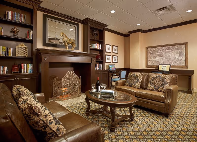 A cozy living room area with two brown leather sofas adorned with patterned cushions, a wooden coffee table with a bottle of wine, two glasses, and books on it. The room features a decorative fireplace with an ornate screen, built-in bookshelves filled with books and decorative items, and two computer desks with chairs along the back wall. The carpet has a patterned design, and the walls are decorated with framed artwork.